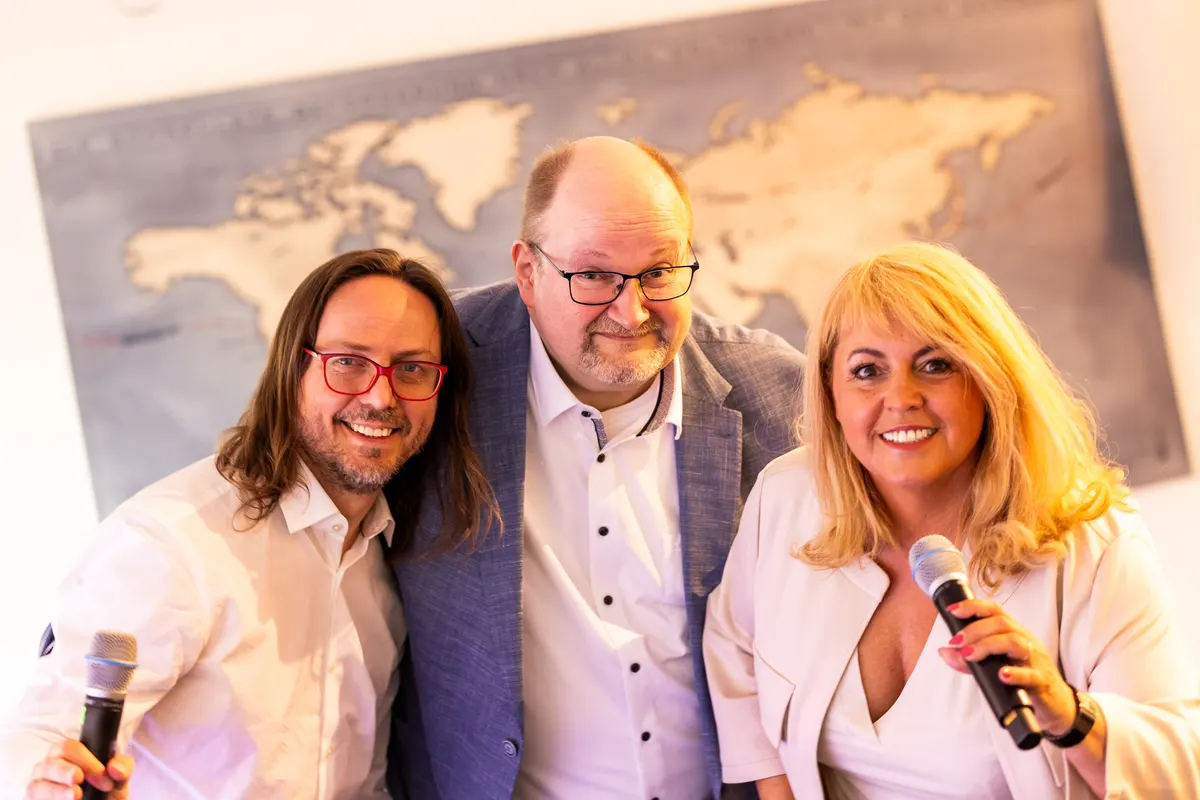 Claudia Conen mit Mikrofon und zwei Gästen vor einer Weltkarte