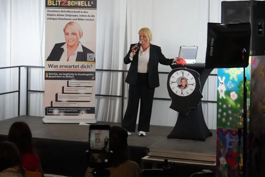 Claudia Conen mit Mikrofon auf einer kleinen Bühne, Publikum von hinten